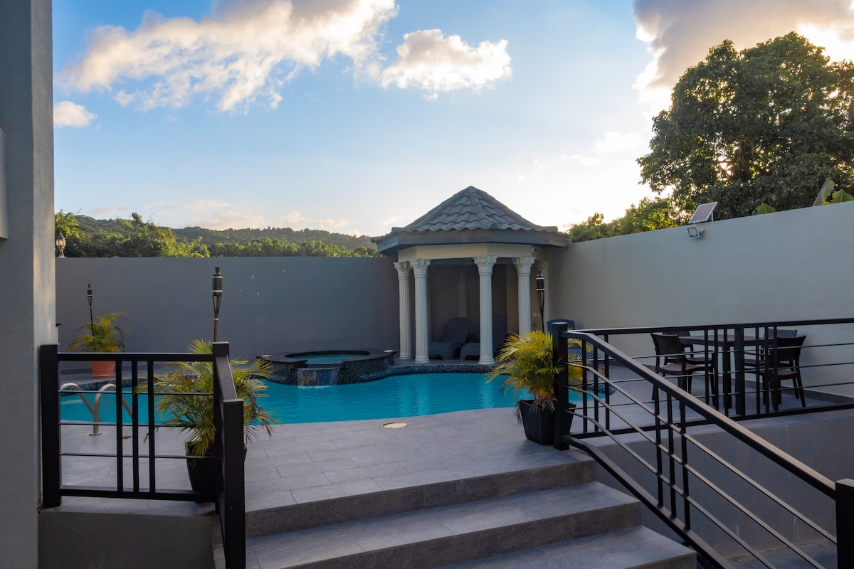 A secluded outdoor area features a tranquil swimming pool, complemented by a gazebo and lounge seating. Surrounding greenery enhances the area, while grey stone steps lead down to the poolside. A dining table with chairs is positioned nearby, providing a space for relaxation and enjoyment.