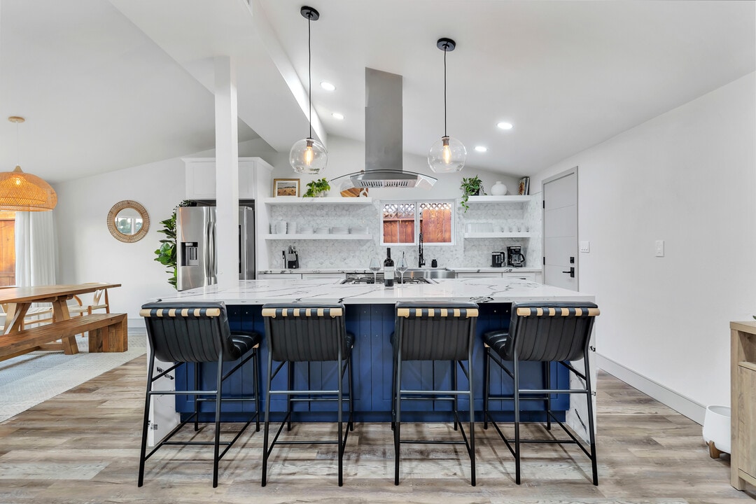 A modern kitchen features a large island with four black barstools. The stainless steel appliances and stylish cabinetry are complemented by a sleek countertop. Natural light fills the space, highlighting the open layout that connects to a dining area in the background.