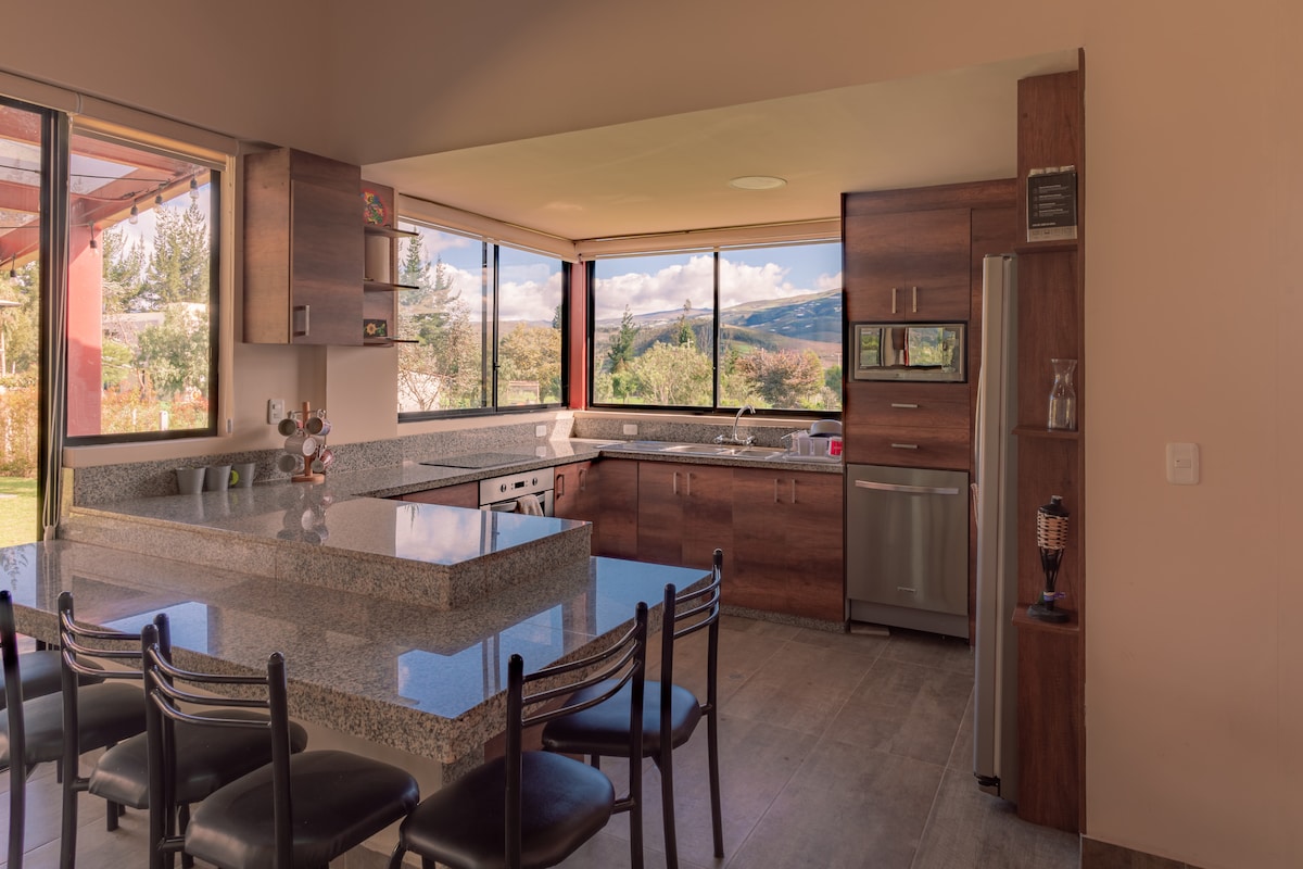 A modern kitchen features an extensive granite countertop with bar seating for six. Large windows provide natural light and views of the surrounding landscape, while stainless steel appliances, including a refrigerator, oven, and microwave, are visible in the cabinetry.