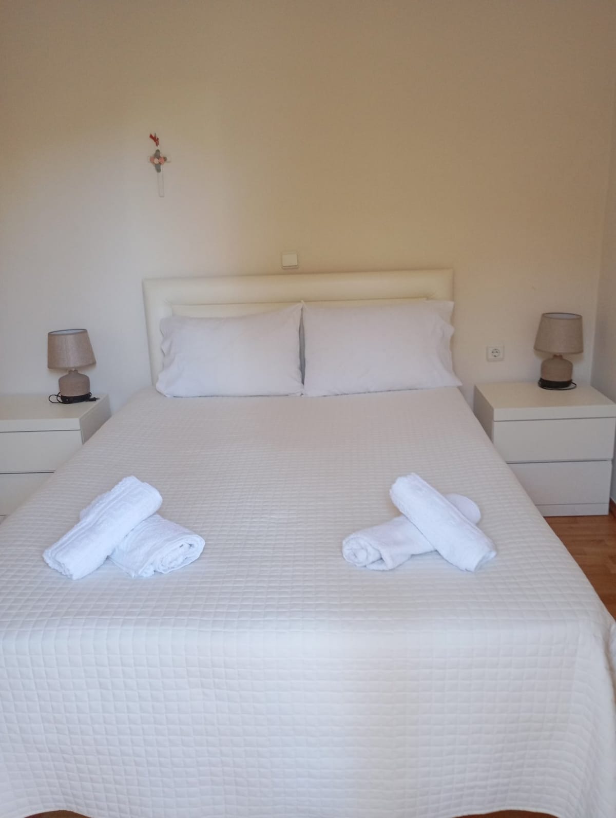 A spacious bed with crisp white linens is centered in the room. Two bedside tables, each with a lamp, are positioned on either side. Fresh towels are neatly arranged on the bed, enhancing the room's clean and welcoming atmosphere.