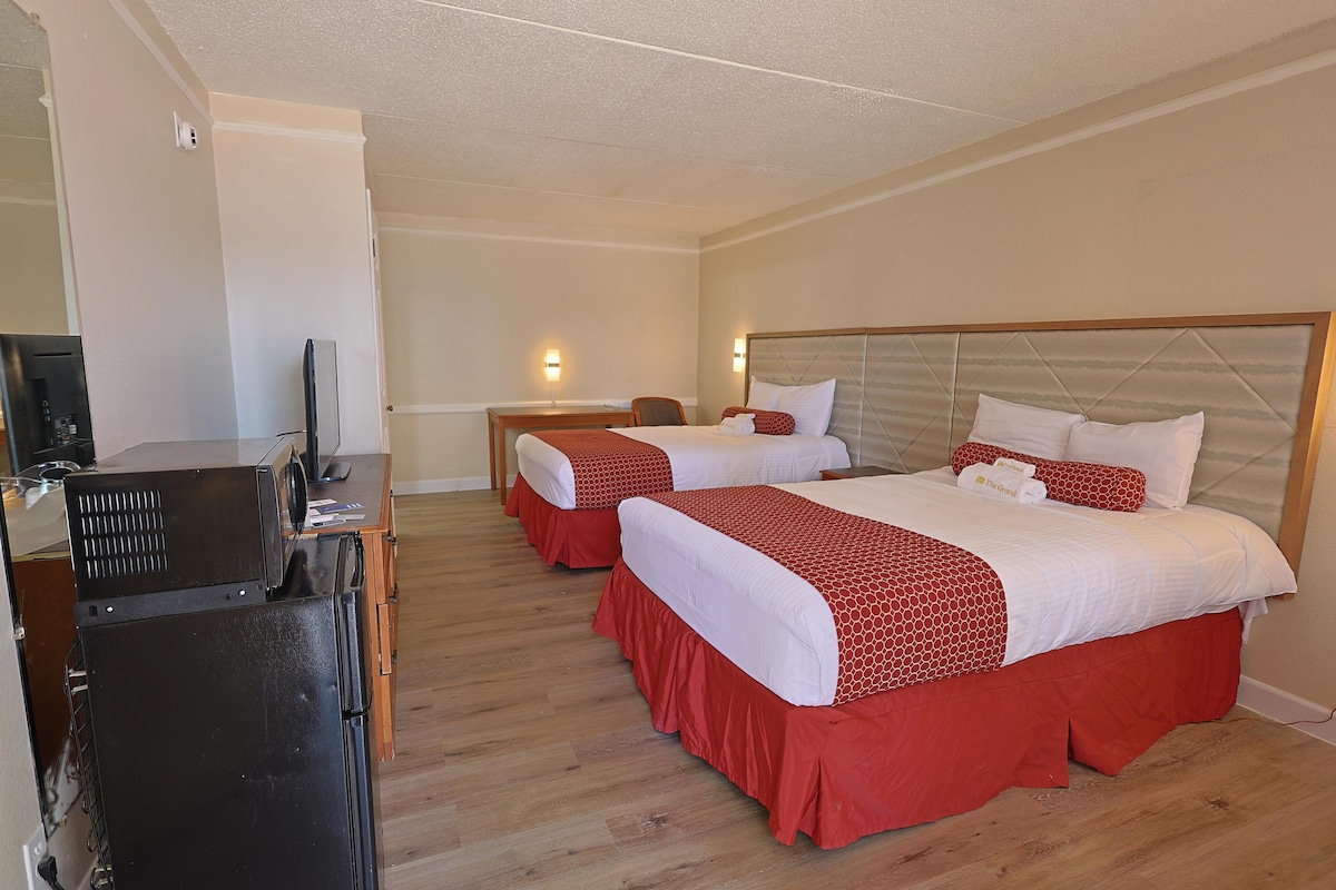 A spacious guest room is equipped with two neatly made beds, each topped with white linens and decorative red throws. A wooden dresser and a compact refrigerator are positioned against the wall, while a wall-mounted TV is visible for entertainment.