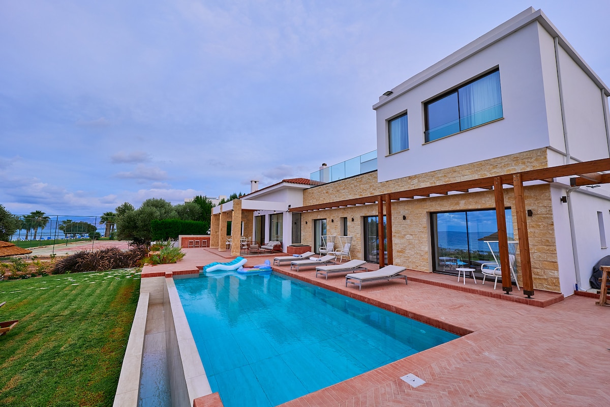 The exterior of the villa is displayed, featuring a modern design with stone and white walls. A tranquil pool runs alongside the patio, surrounded by sun loungers. An expansive yard with green grass leads towards the beachfront, with views of the sea visible in the background.