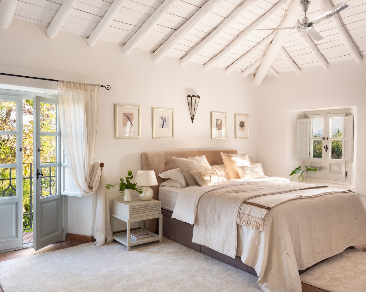 A light-filled bedroom features a spacious bed dressed in neutral tones, complemented by decorative wall art. Large windows offer scenic views and allow natural light to illuminate the room. Soft furnishings and plants add a touch of warmth to the serene atmosphere.