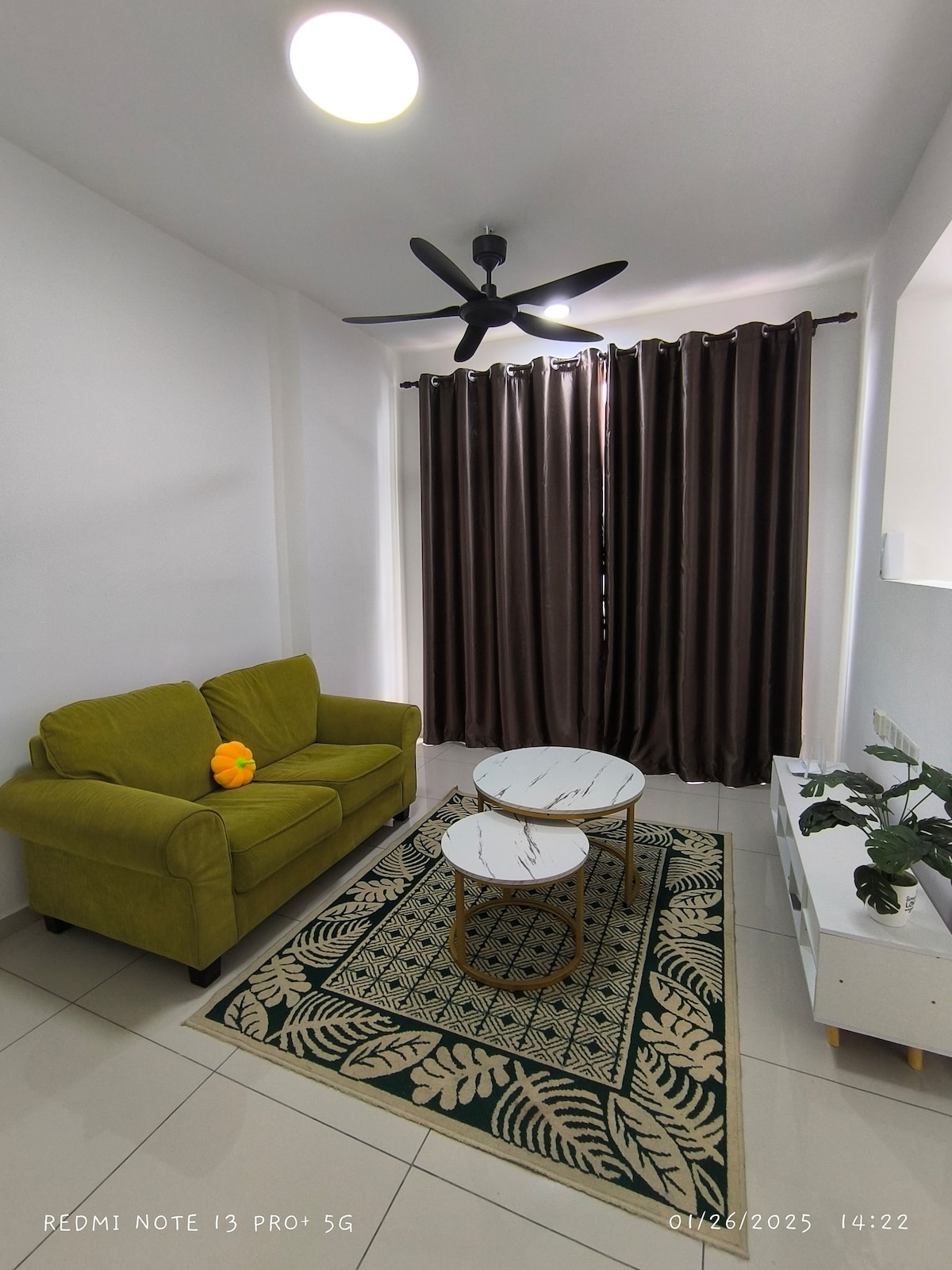 A cozy living area features a green sofa accompanied by two round coffee tables on a patterned rug. Natural light filters through dark curtains, and a ceiling fan enhances the air circulation in the space.