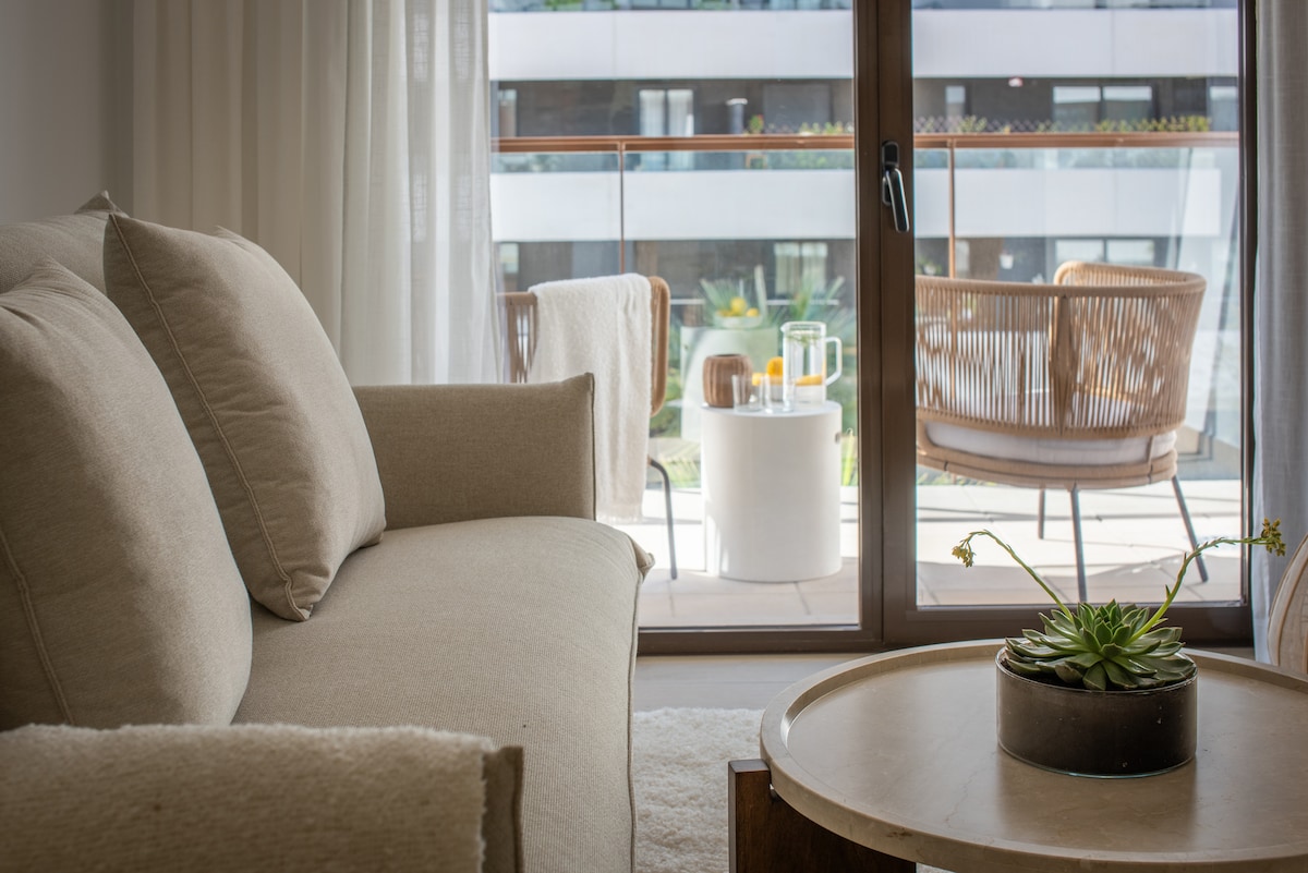 A spacious living area is presented with a neutral-colored sofa, positioned near a large glass door that leads to a private terrace. The terrace features outdoor seating with stylish chairs and a small table, surrounded by greenery, enhancing the inviting atmosphere.