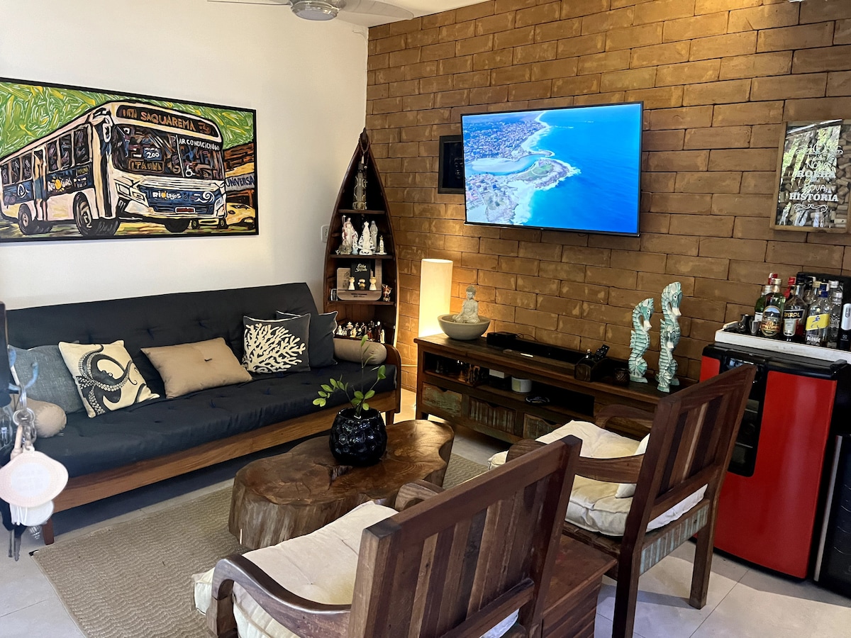 An inviting living area features a dark couch adorned with decorative cushions, complemented by wooden chairs. A large flat-screen TV is mounted on a textured wall, while a rustic coffee table adds character. Decorative accents and plants provide a touch of nature to the space.