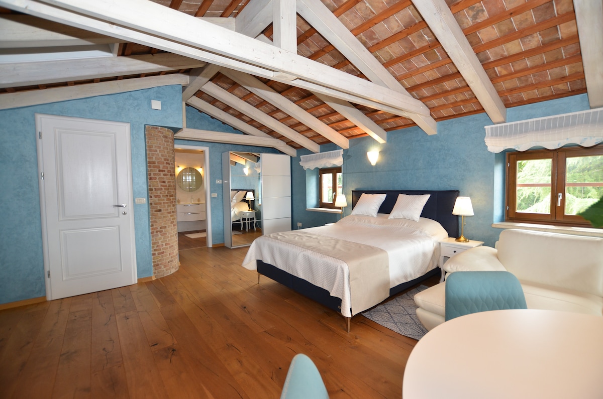 A spacious bedroom features a large bed with a light-colored headboard, surrounded by rustic wooden beams and a warm wood floor. Soft blue walls complement natural light coming through windows, while a cozy seating area with white furniture and a round table sits nearby.