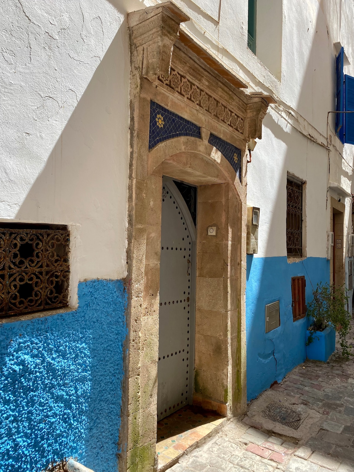 An intricately designed entrance showcases traditional architecture with stone detailing and decorative blue tiles. The door, framed by an arched stone structure, stands against a backdrop of white and blue walls, enhancing the charm of the narrow street.