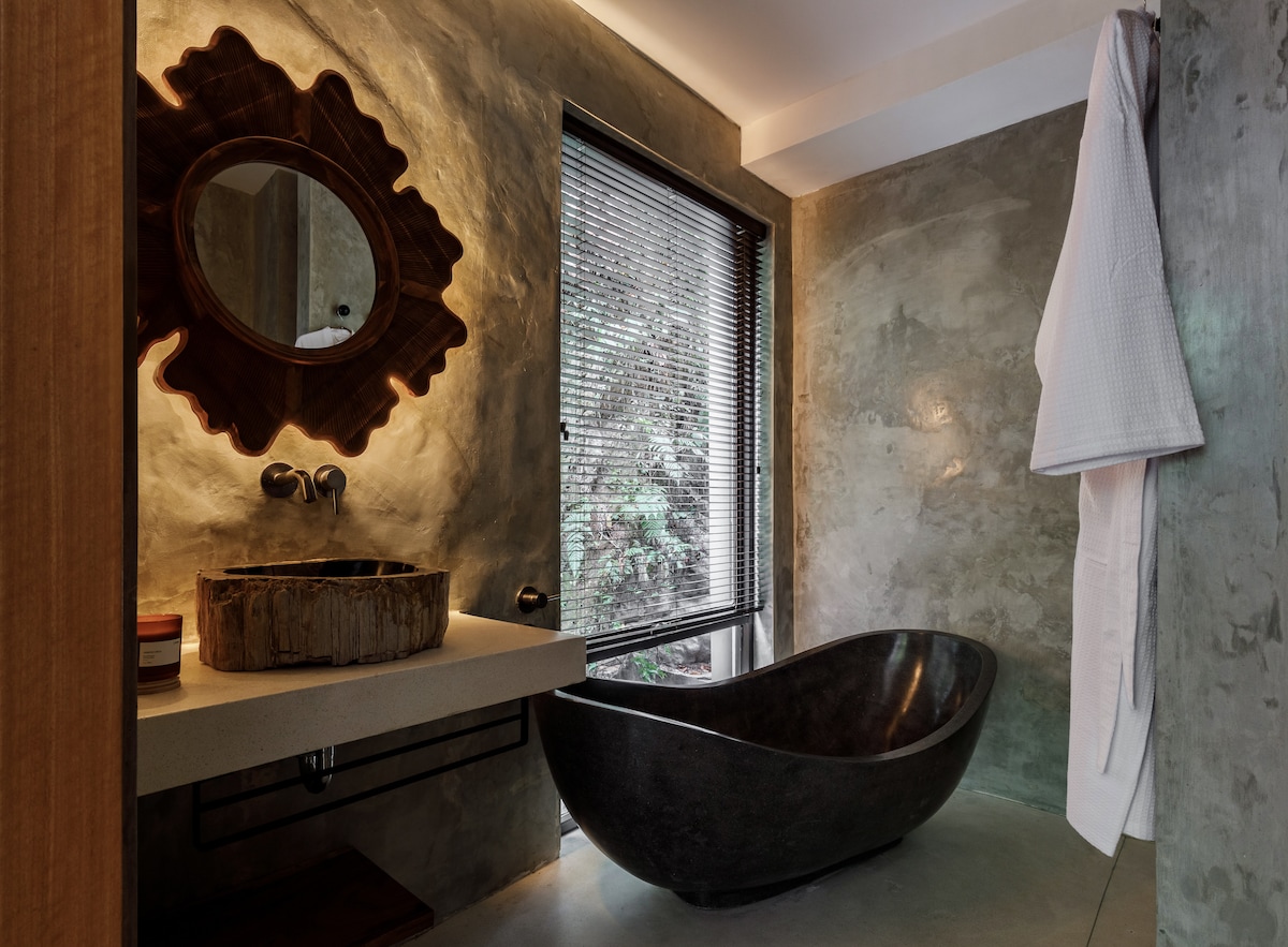 A modern bathroom features a smooth stone freestanding tub beside a large window offering views of greenery. A unique mirror complements the artistic sink crafted from natural wood, while soft ambient lighting enhances the minimalist decor.