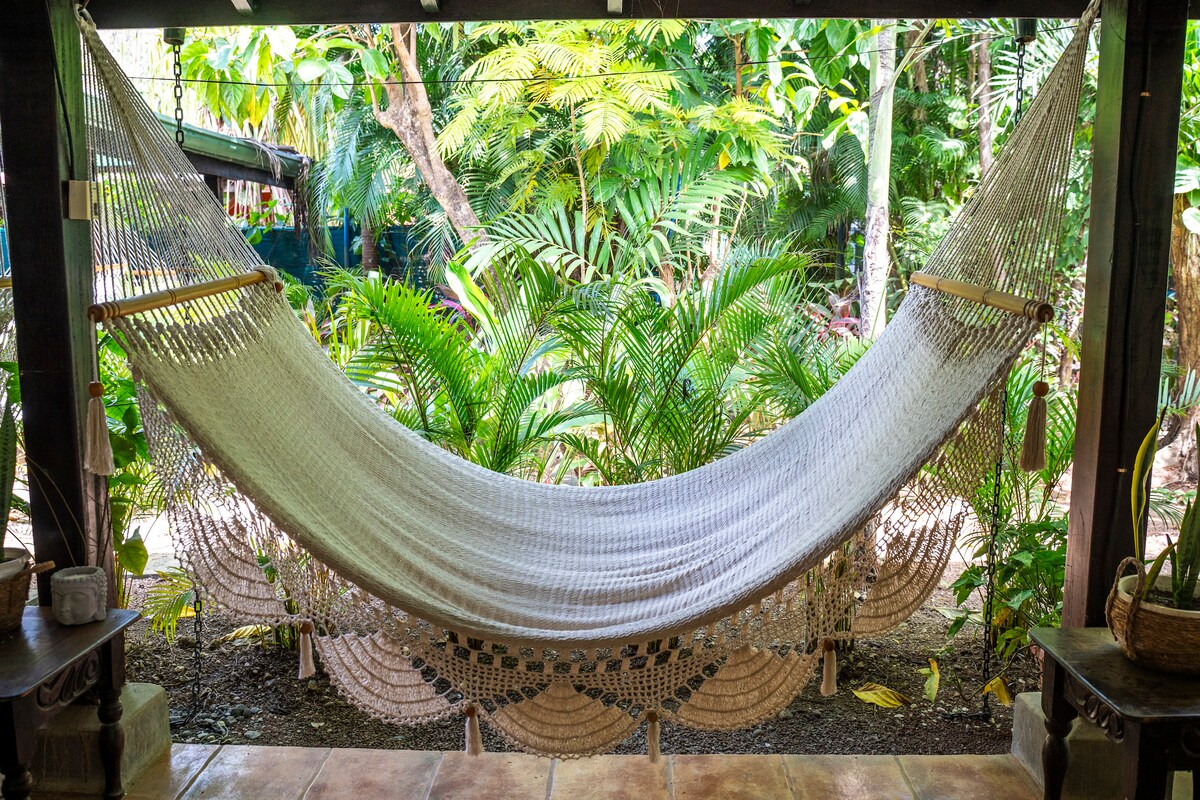A spacious hammock is suspended under a covered area, surrounded by lush tropical plants. The natural setting provides a serene backdrop, with various shades of green creating a peaceful atmosphere. The ground is paved with warm-toned tiles, enhancing the inviting space.