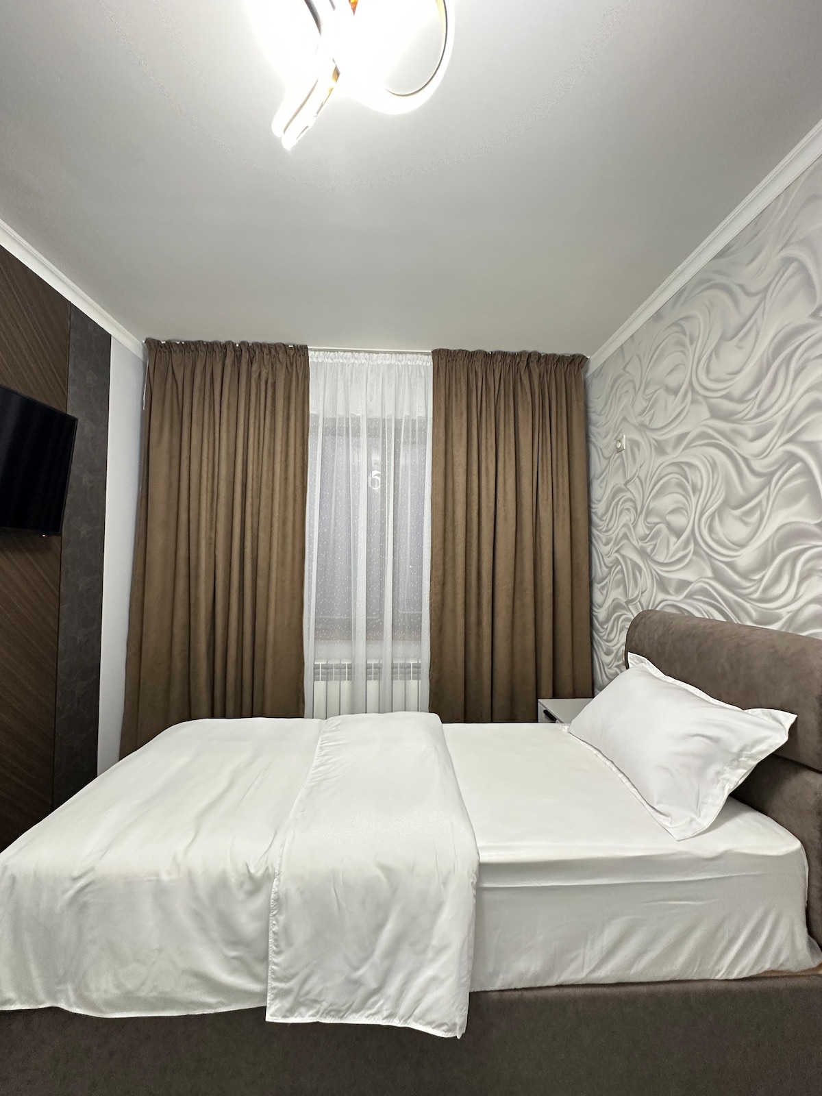 A neatly arranged sleeping area features a spacious bed dressed in crisp white linens. Walls are adorned with textured panels, while long, neutral-colored curtains frame the window. A small bedside table is positioned beside the bed, and a television is mounted on the wall.