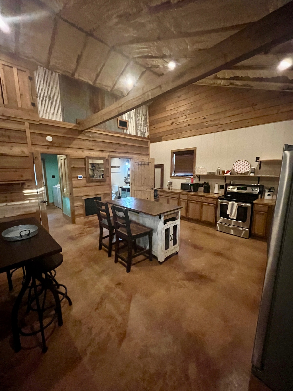 The kitchen area features wood-paneled walls and a high ceiling, creating an open atmosphere. A spacious island with seating for four is visible, complemented by modern appliances, including a refrigerator and oven. Natural light is welcomed through several windows.