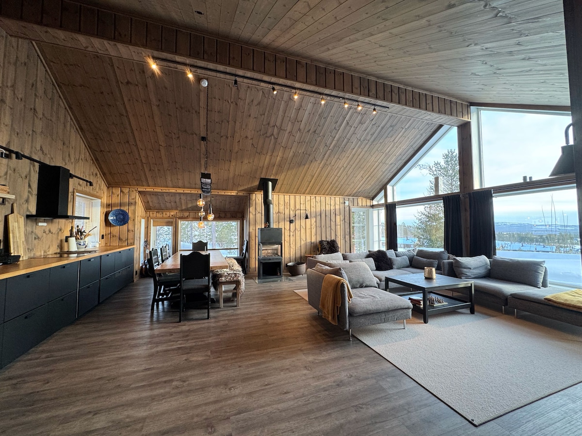 The spacious living area features a high, slanted wooden ceiling with recessed lighting. A large seating arrangement includes multiple sofas and cushions, arranged around a central coffee table. A dining area with a long table is visible, alongside large windows offering scenic views.