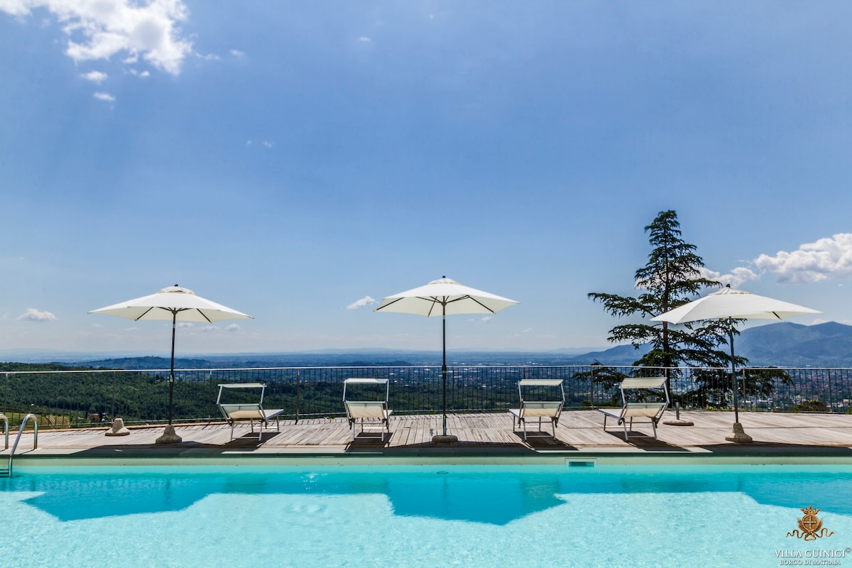 A tranquil swimming pool is framed by a sun deck featuring four loungers and large umbrellas. Rolling hills and a clear blue sky create a serene backdrop, enhancing the inviting atmosphere of relaxation and comfort.