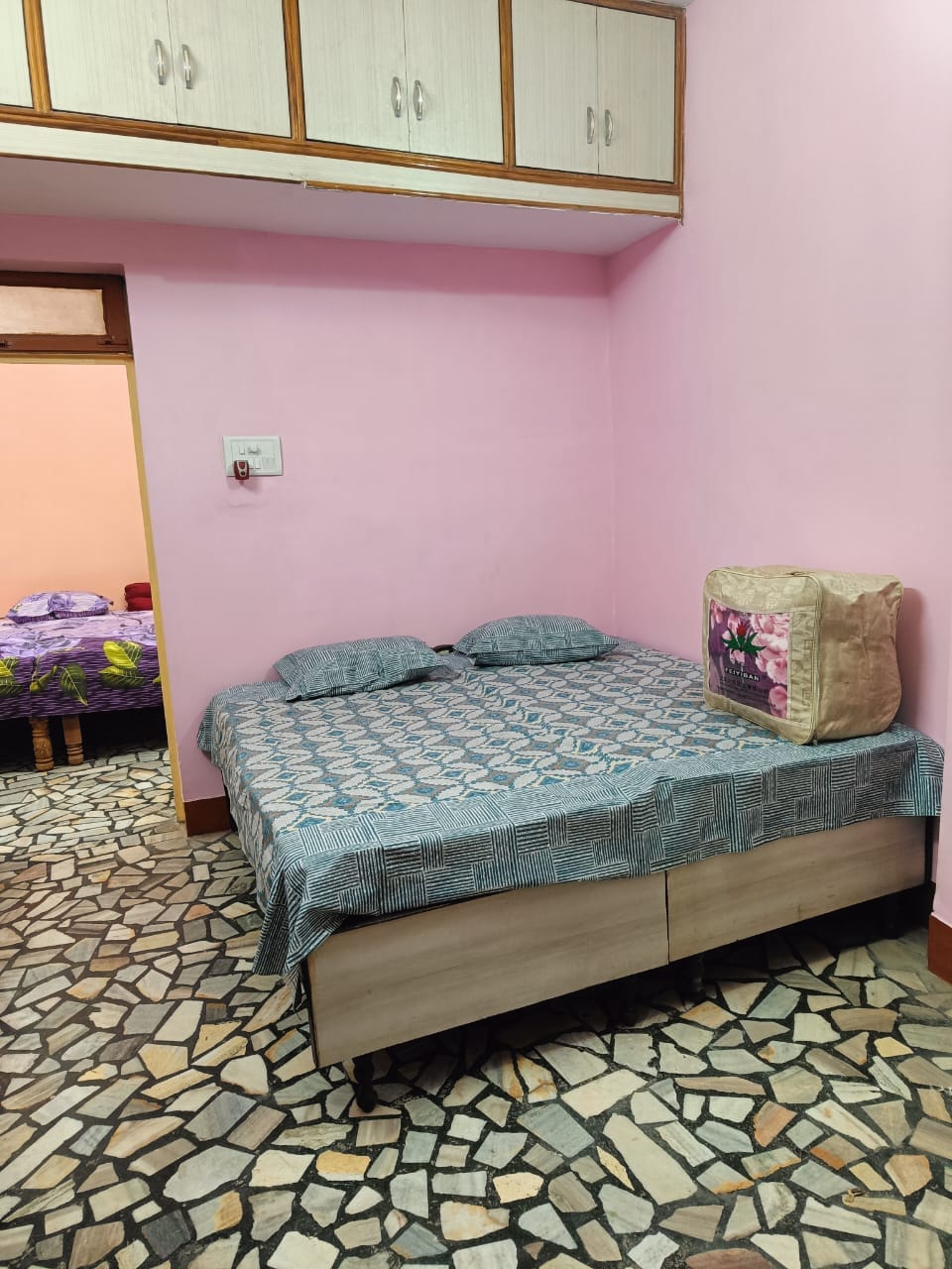 A neatly arranged sleeping area features a spacious bed covered with a patterned blue quilt and two pillows. The walls are painted in a soft pink hue, complemented by a decorative cushion on the bedside. A unique mosaic floor adds character to the room.