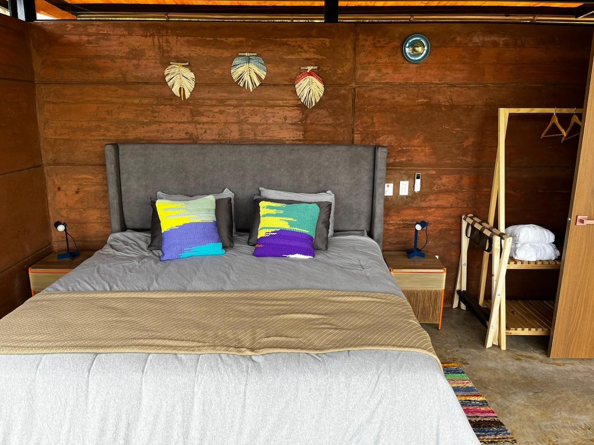A spacious sleeping area showcases a large bed with colorful decorative pillows. Nightstands with lamps are positioned on either side. Natural wood walls contribute to a cozy ambiance, while a wardrobe and hanging space provide convenient storage options.