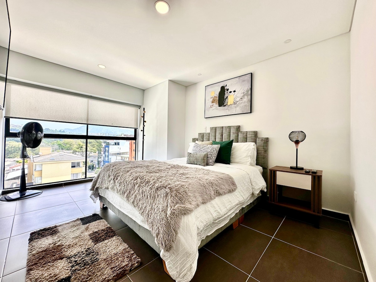 A bright bedroom features a spacious bed with a textured headboard and soft bedding. A small bedside table with a lamp is positioned beside the bed. Natural light filters through a large window, offering views of the surrounding landscape. A cozy rug adds warmth to the tile floor.