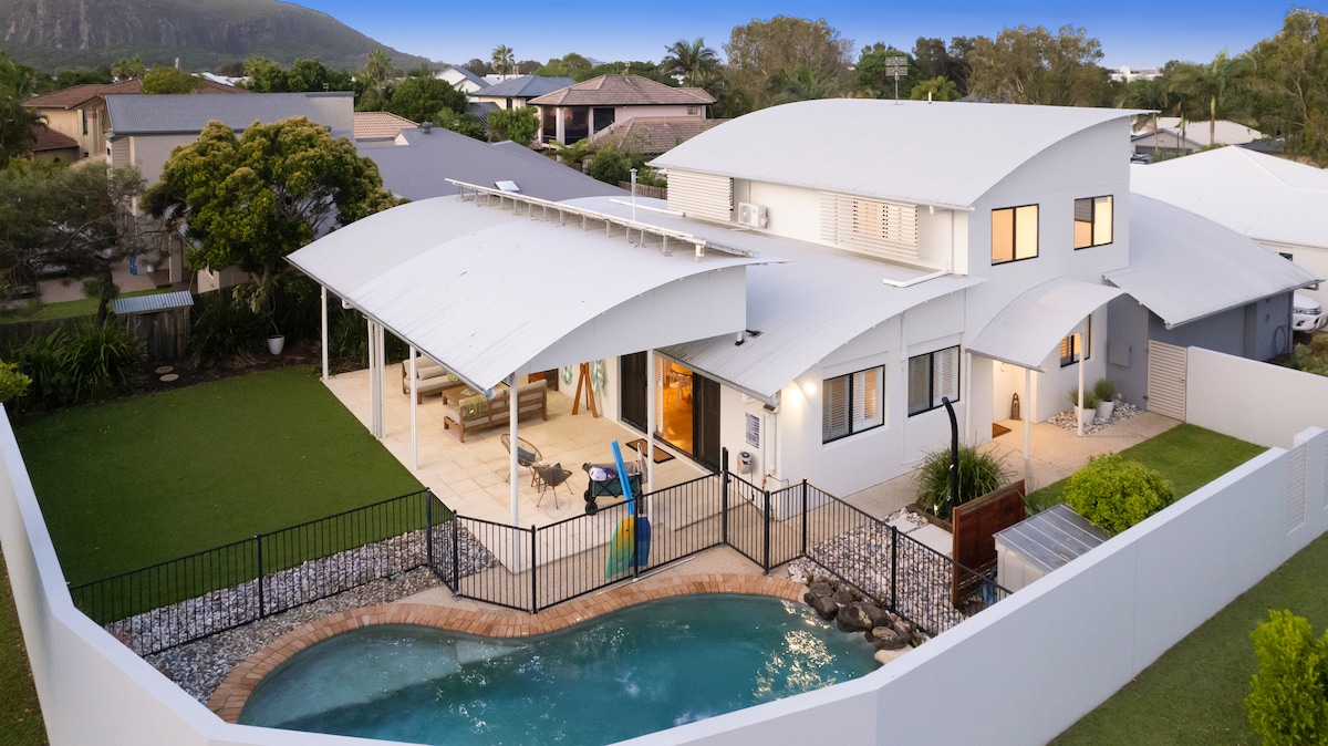 An aerial view captures a spacious coastal retreat, featuring a sparkling pool surrounded by secure fencing. Lush green lawns and a large covered alfresco area are visible, along with a modern structure showcasing multiple rooflines and large windows that invite natural light.