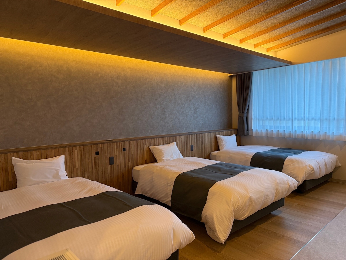 A spacious guest room features three semi-double beds with white bedding and dark green throws. Warm wooden accents create a calming environment. Natural light filters through sheer curtains, enhancing the inviting and functional space.