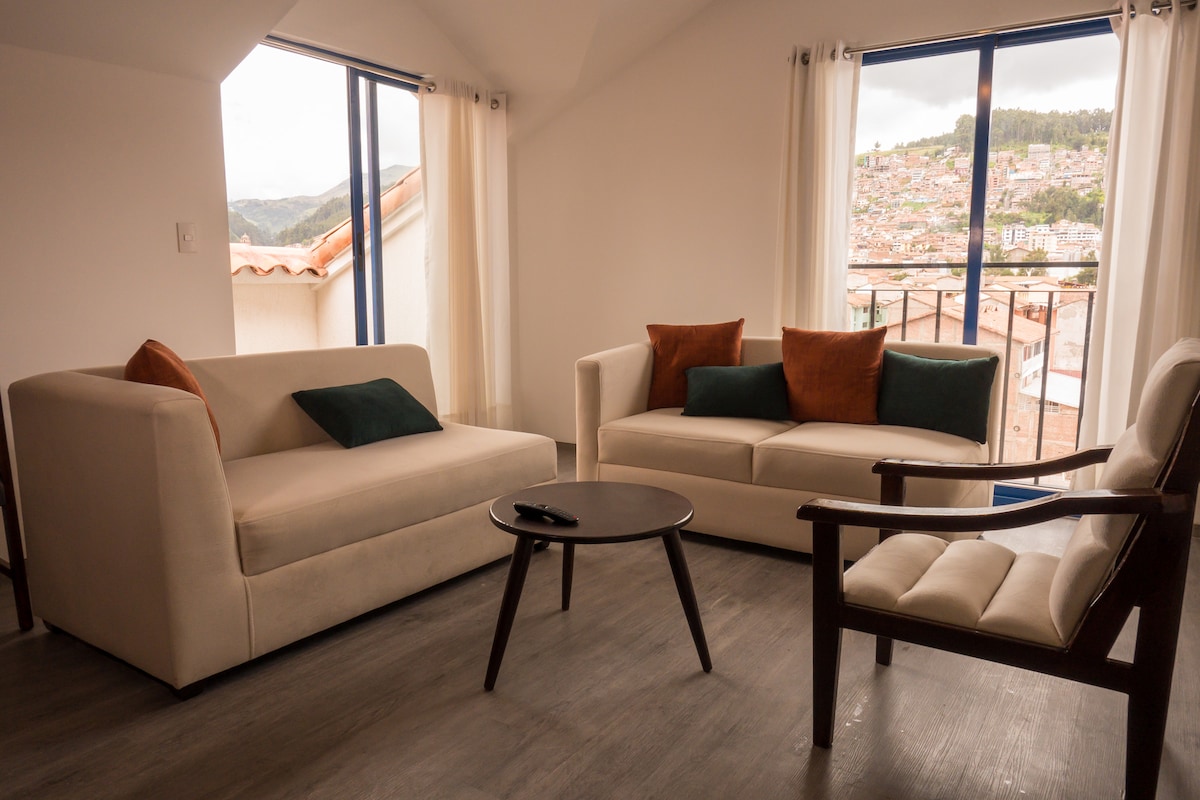 A seating area features a light-colored sofa and a matching loveseat, complemented by a wooden chair. Decorative cushions in earthy tones are arranged on the sofas. A round coffee table sits at the center, with large windows providing views of the surrounding landscape.
