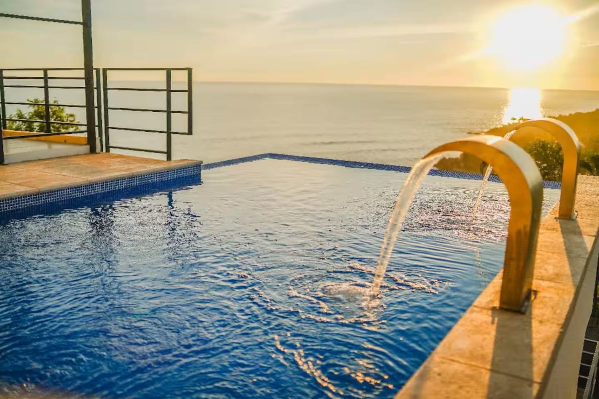 An infinity pool extends toward the horizon, presenting a seamless view of the ocean at sunset. Water flows gently from two elegant spouts into the vibrant blue water, reflecting the warm colors of the sky.