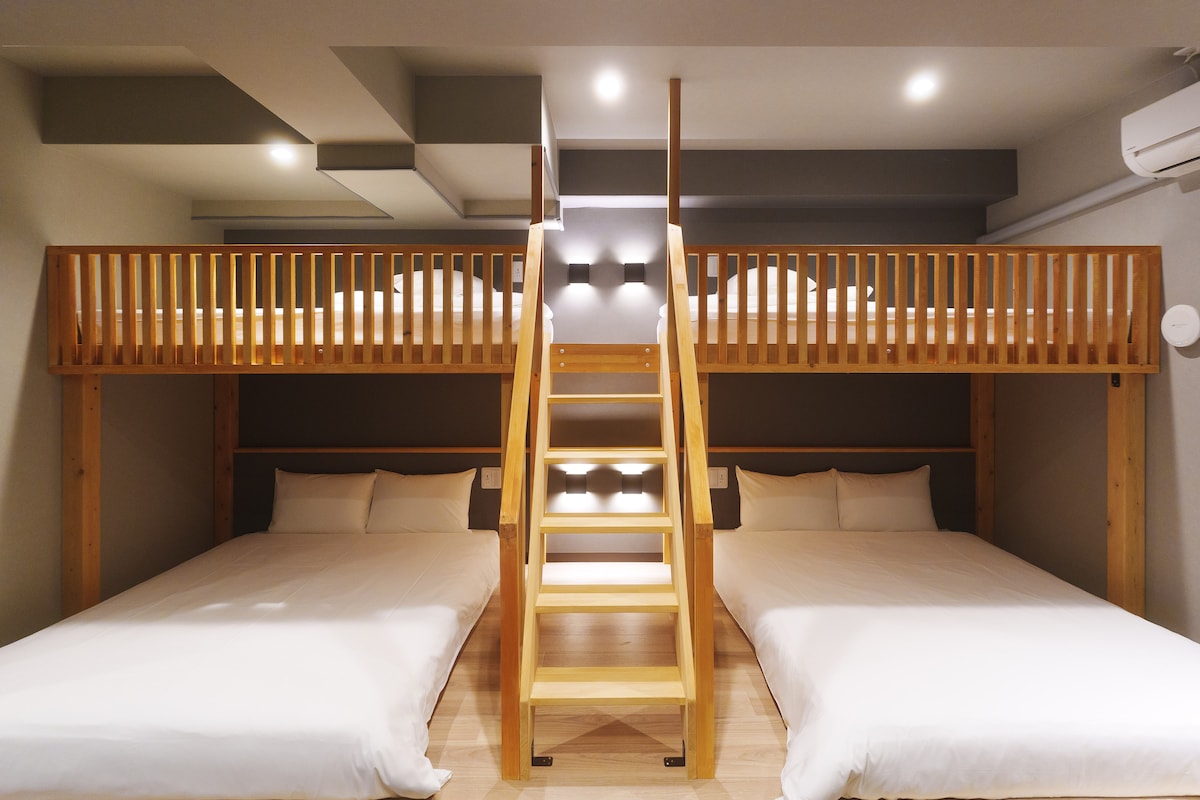 A spacious room features two large beds on the lower level, while a wooden loft bed is situated above, accessed by a sturdy ladder. Soft lighting enhances the modern design, and the neutral color palette creates a calming environment.