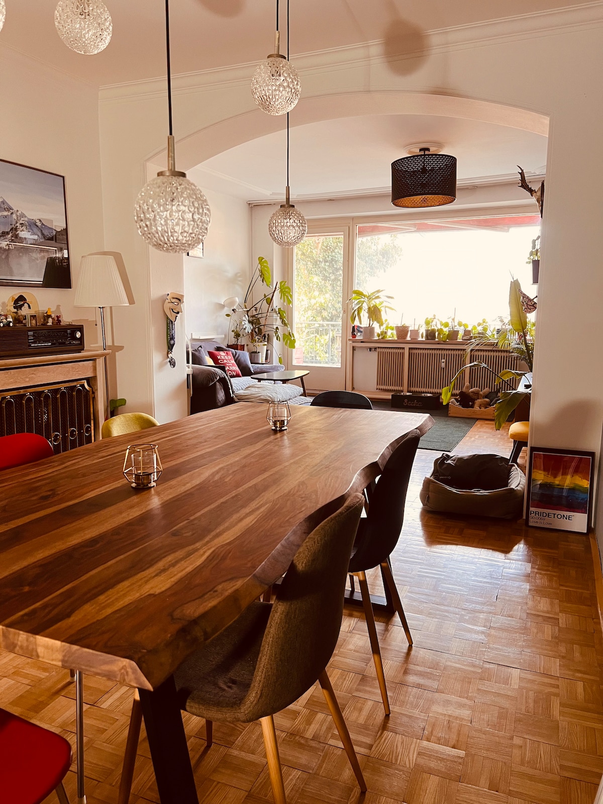 A spacious dining area is highlighted by a large wooden table surrounded by various chairs in different colors. Pendant lights hang above the table, providing soft illumination. A large window allows natural light to fill the room, enhancing the view of plants and seating in the background.
