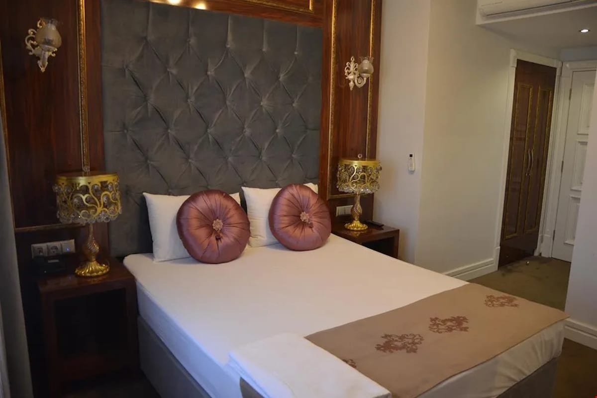 The room features a stylish bed with an elegant tufted headboard. Pink decorative pillows are placed against a gray backdrop. Two bedside lamps with ornate bases provide soft lighting, enhancing the modern design of the space.