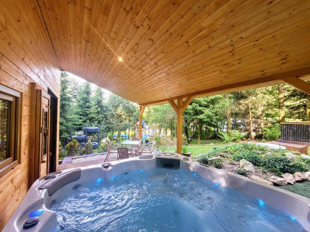 An outdoor hot tub is situated under a wooden gazebo, surrounded by lush greenery and tall trees. Soft lighting illuminates the area, providing a relaxing ambiance. Nearby seating arrangements are visible, enhancing the space for socializing or enjoying the serene view.