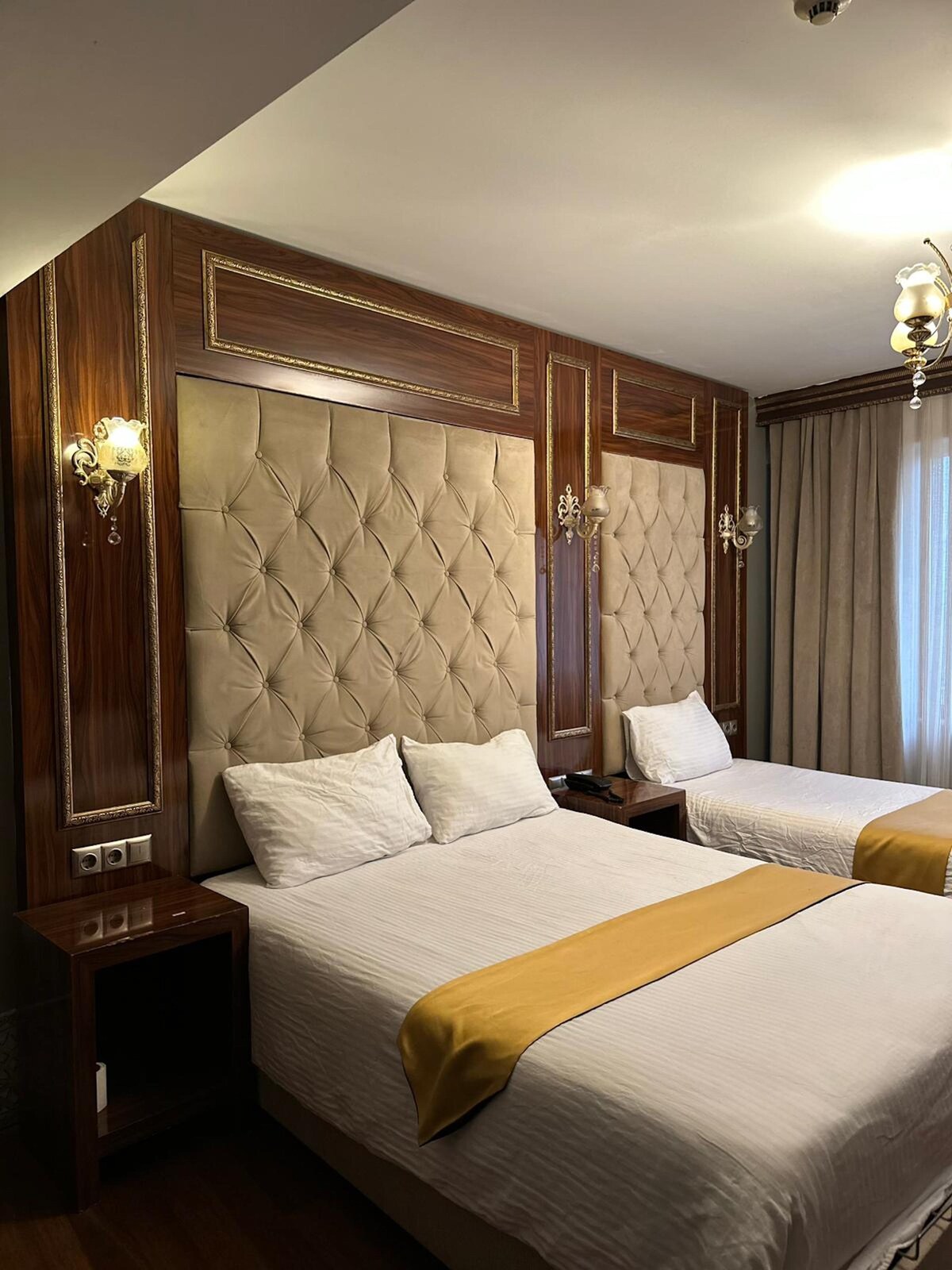 A modern guestroom features two spacious beds with upholstered headboards, accented by elegant wall sconces. Light-colored linens are complemented by a gold decorative throw, and soft natural light filters through the floor-to-ceiling curtains, enhancing the room's refined design.