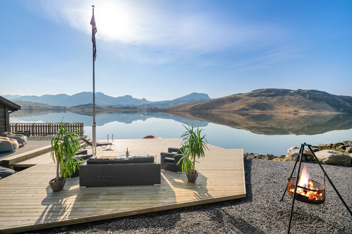A wooden deck extends toward a calm body of water, featuring a cozy seating area with dark outdoor furniture. Potted plants add a touch of greenery, while a flagpole stands nearby. A fire pit offers a warm focal point against a backdrop of scenic hills.
