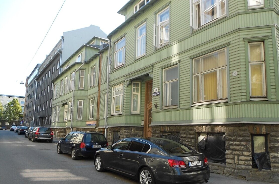 The exterior of a green wooden building is depicted, showcasing multiple windows with light flowing through the panes. Cars are parked along the street, and the building features decorative woodwork that highlights its architectural details.