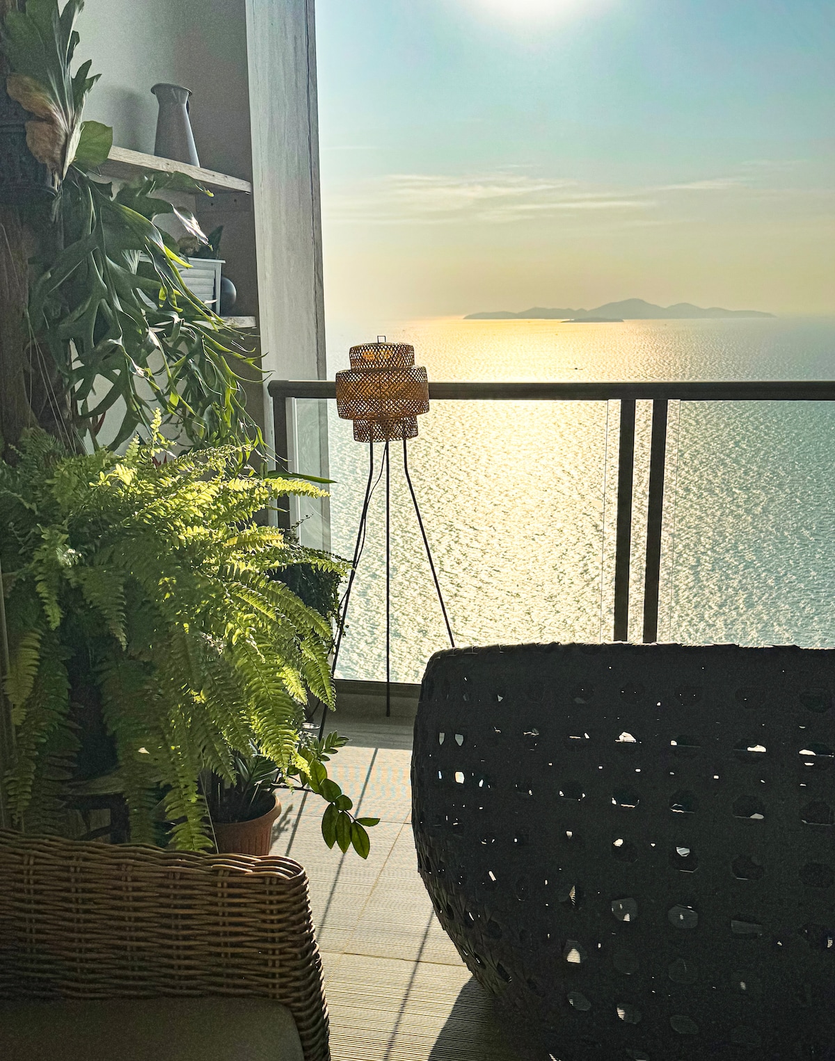 A cozy terrace corner features a decorative lamp on a slender stand, surrounded by lush green ferns. The tranquil ocean view can be seen in the background, with the sun reflecting brightly off the water, creating a serene atmosphere.