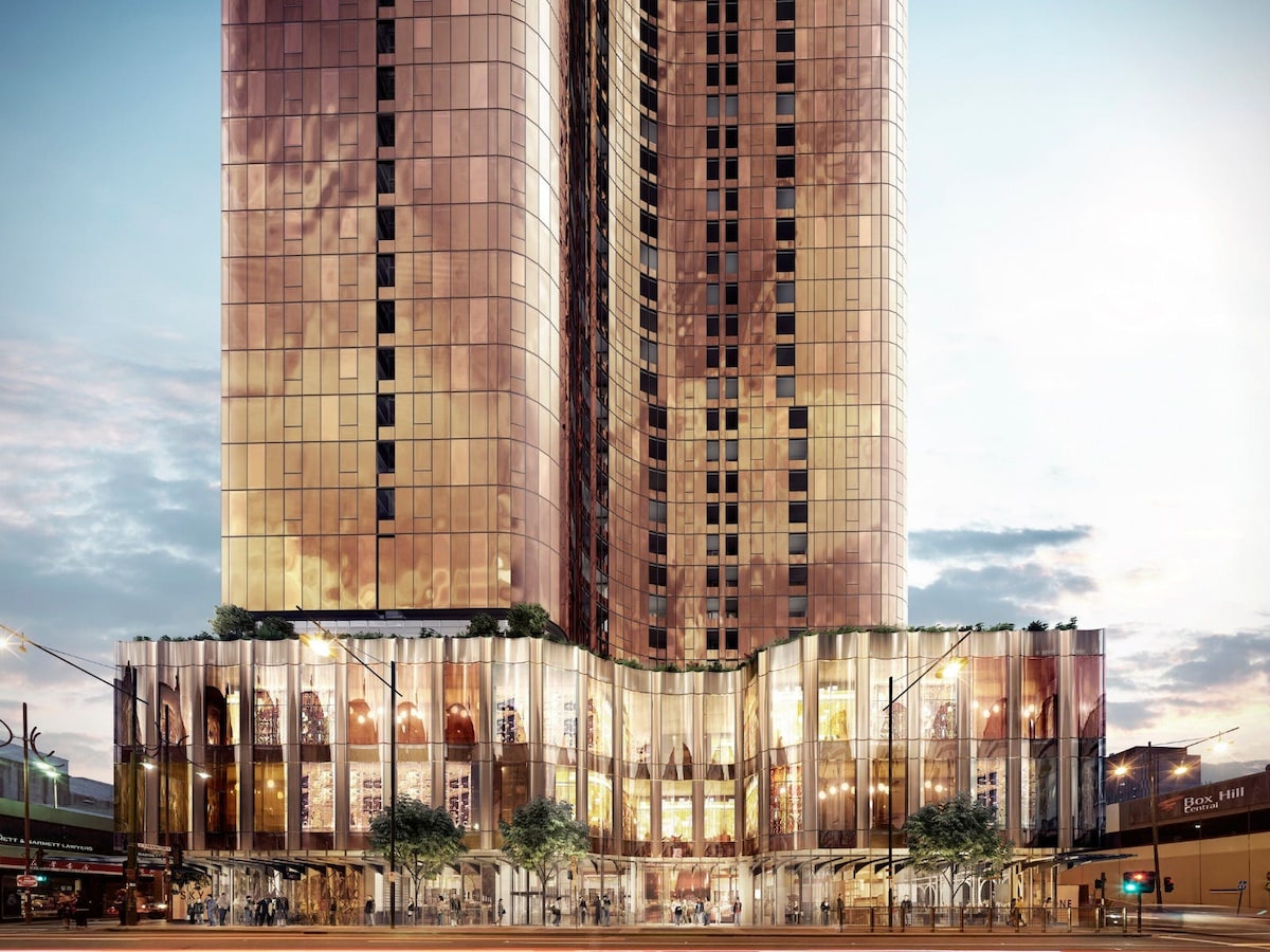A modern high-rise building features a sleek glass façade that reflects the surrounding environment. The ground level showcases large windows, providing a view into retail spaces. Lush trees are positioned at the base, enhancing the urban landscape.