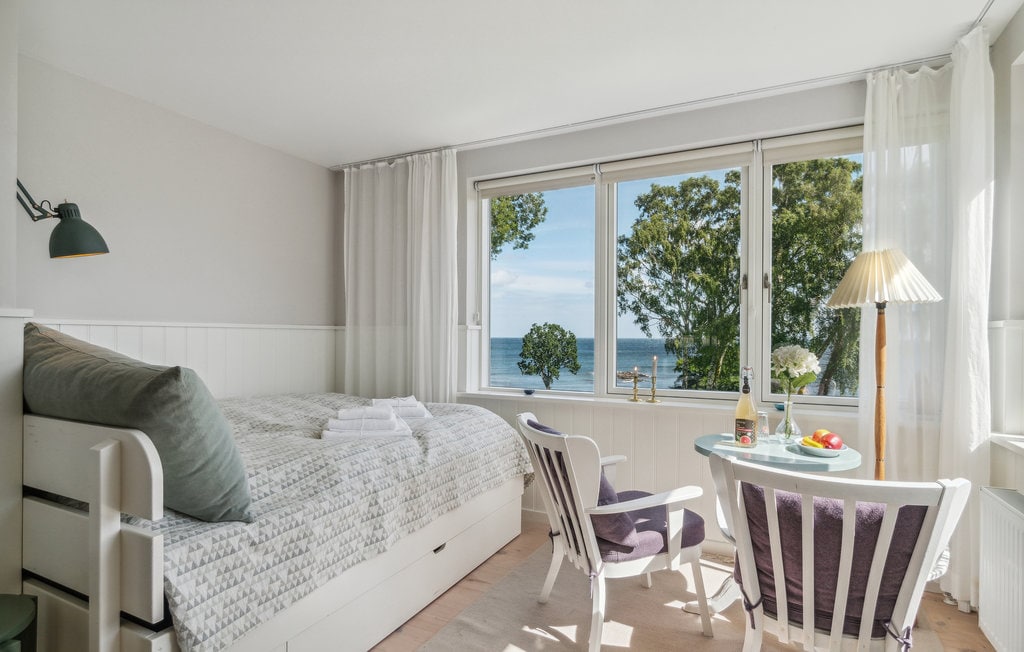 A bright bedroom showcases a spacious bed dressed in neutral linens, accompanied by folded towels. A small round table with two chairs is positioned near large windows that provide a view of the sea and greenery outside. Soft curtains allow natural light to fill the room.