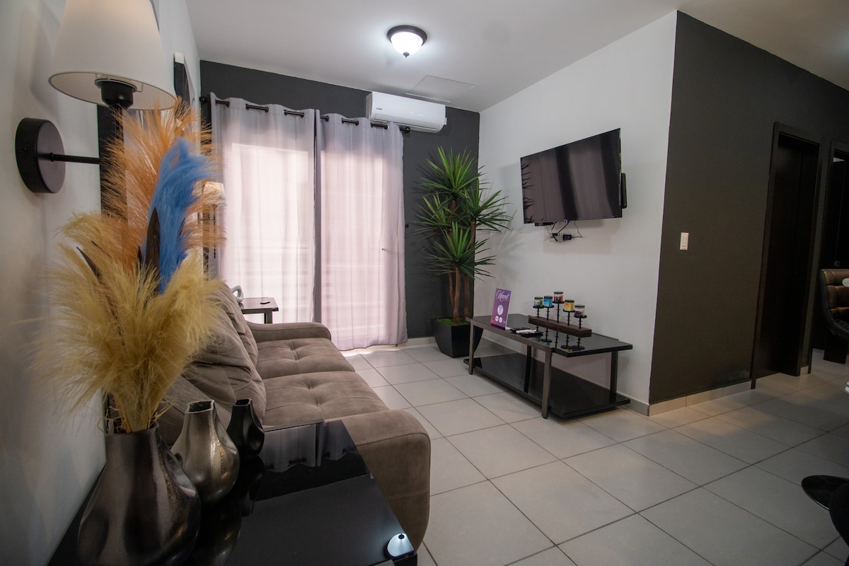 A modern living area features a comfortable, grey sofa paired with a sleek, dark coffee table. A large television is mounted on the wall across from the sofa. Natural light filters through sheer curtains, and a tall plant adds a touch of greenery to the space.