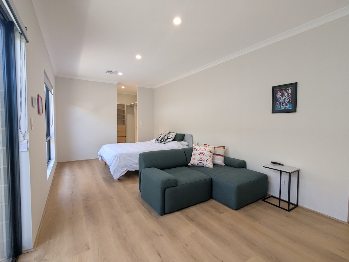 A spacious studio interior features a queen-size bed with crisp linens and a cozy lounge area, which includes a sectional sofa. A small side table is situated beside the sofa. Natural light fills the room through large windows, enhancing the wood-style flooring.