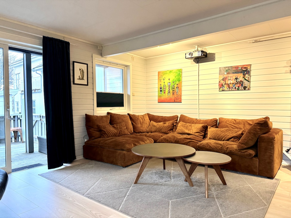 A spacious living area features a large, soft brown sofa complementing two round coffee tables. Light art pieces adorn the white walls, while natural light enters through sliding glass doors. A projector is mounted on the ceiling, enhancing the functional atmosphere.