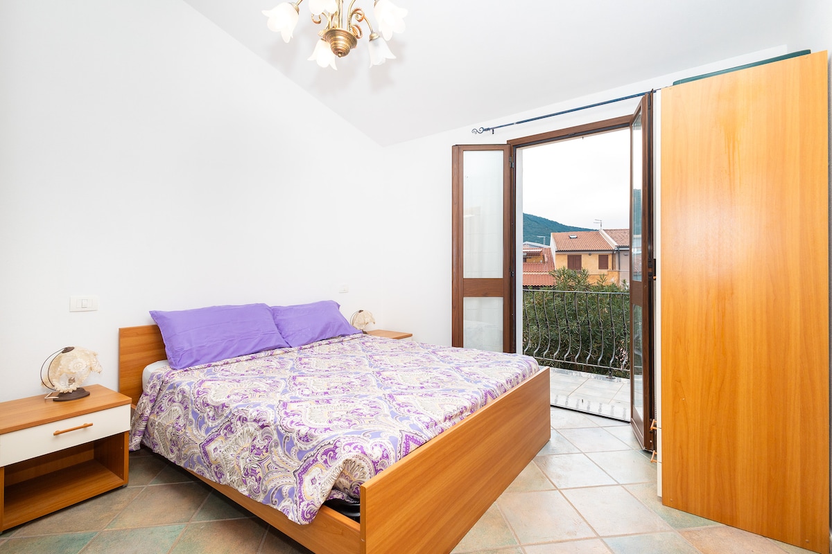 A well-lit bedroom features a spacious bed with a patterned purple and white blanket. Natural light filters through a large open window, which offers a view of the surrounding area. A wooden nightstand sits beside the bed, enhancing the room's simplicity.