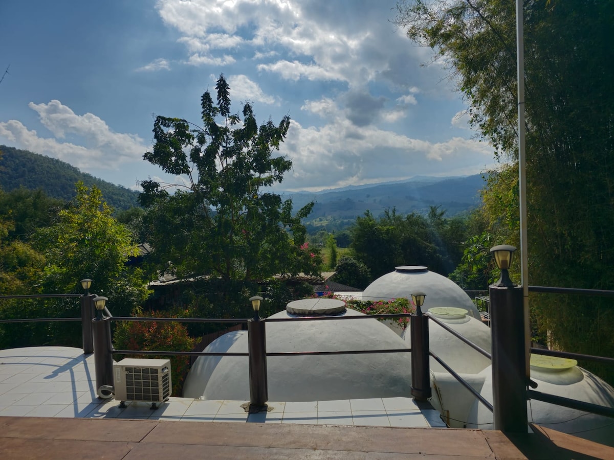 A scenic view showcases rolling hills under a partly cloudy sky. Lush greenery surrounds domed structures, while a railing adorned with lights lines the terrace. The landscape is dotted with trees, providing a serene atmosphere and a sense of seclusion.