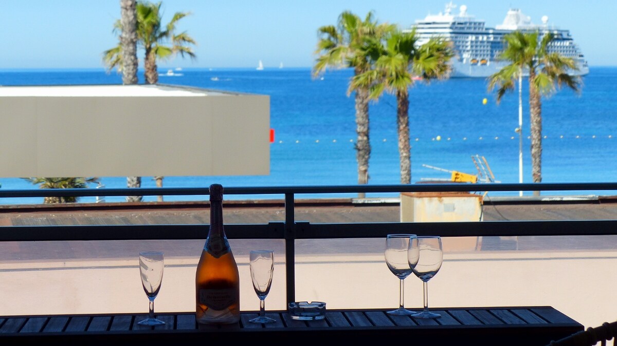 A balcony view features a clear blue sea and palm trees swaying gently in the breeze. A bottle of champagne stands on a wooden table, accompanied by four empty glasses, ready for a toast against the backdrop of a distant cruise ship.