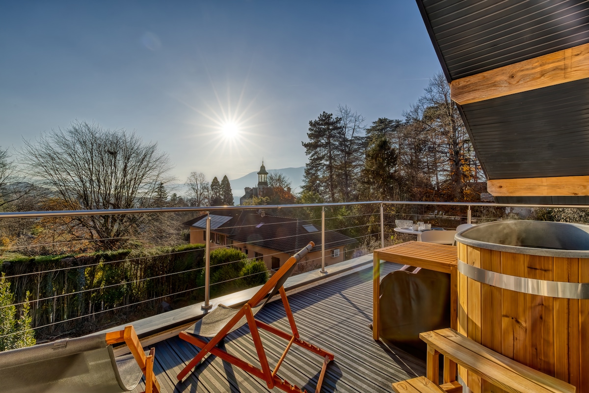 A sunlit rooftop terrace is presented, featuring a private Nordic bath and a wooden lounge chair. Surrounding greenery and distant mountains create a serene backdrop, while a clear blue sky enhances the tranquil atmosphere.