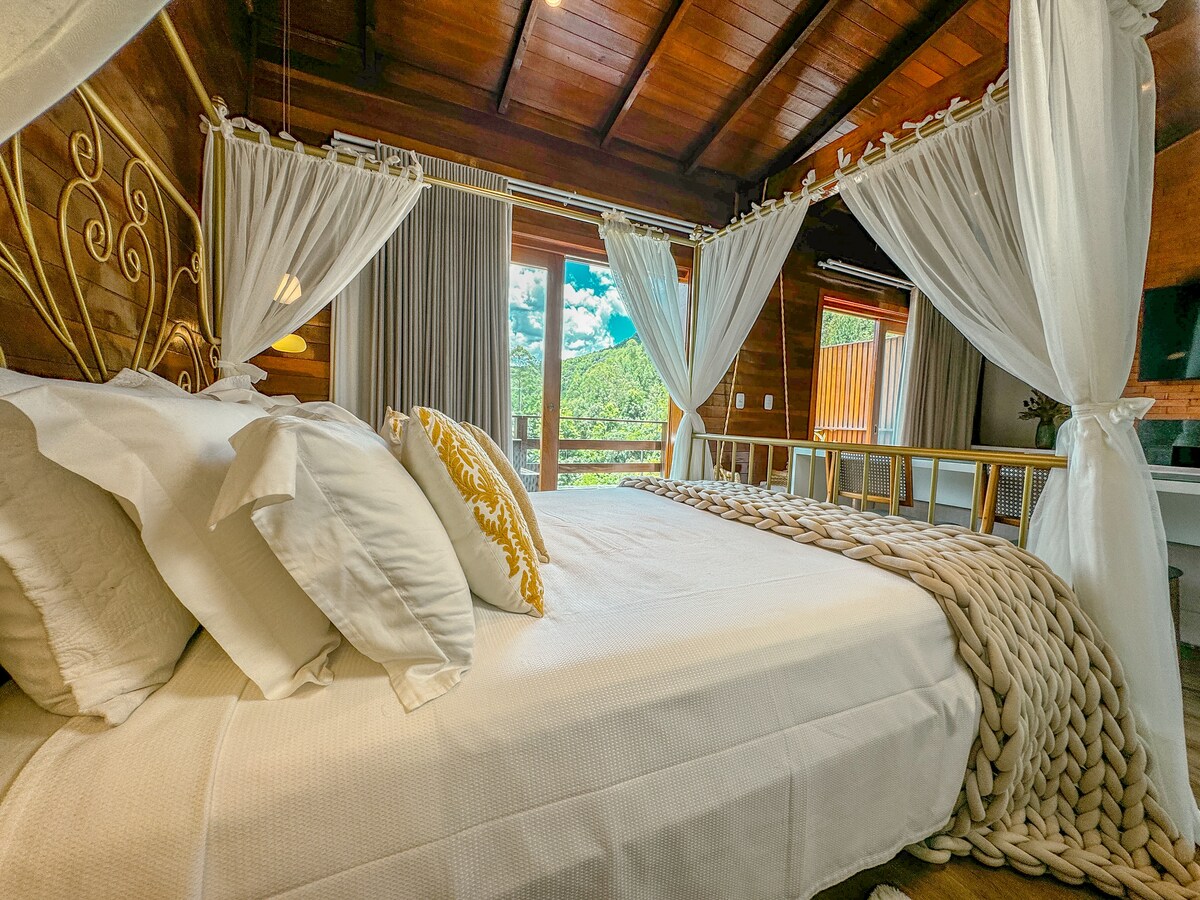 A well-lit bedroom features a spacious bed with decorative pillows and a textured blanket. Soft drapes frame the windows, which offer views of lush greenery. The rustic wooden ceiling adds warmth, and light fills the room, creating a serene ambiance.