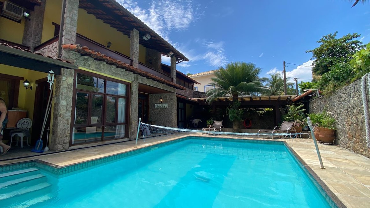 A refreshing outdoor pool is showcased, surrounded by a stone patio. Sun loungers are positioned nearby under shaded areas, and lush greenery is visible in the background. The house's two-story structure features large glass doors, ensuring a connection between indoor and outdoor spaces.