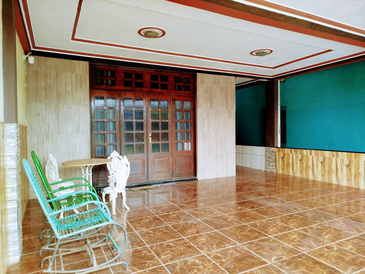 A spacious terrace area is outlined by tiled flooring and features large wooden double doors leading inside. Green rocking chairs and a white table are positioned for relaxation, while walls are adorned with a rich teal color, enhancing the open layout.