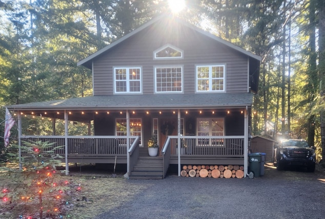 A charming two-story cabin is framed by towering trees, an inviting front porch adorned with lights, and a decorative entrance. A vehicle is parked nearby, complementing the home’s wooden features and natural surroundings.