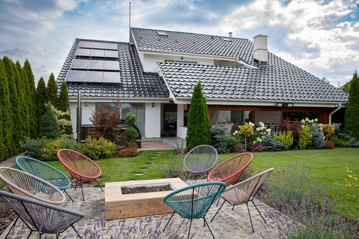 An outdoor seating area features colorful acapulco chairs arranged around a wooden fire pit. Lush greenery and flowering plants surround the space, while a modern home with solar panels is visible in the background, complemented by a serene sky.