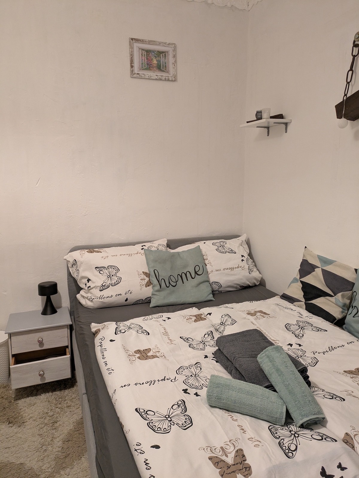A cozy sleeping area is presented with a double bed adorned with decorative pillows and a stylish quilt featuring butterfly prints. A small bedside table with a lamp stands nearby, while a shelf holds decor items above the bed, contributing to a relaxing atmosphere.