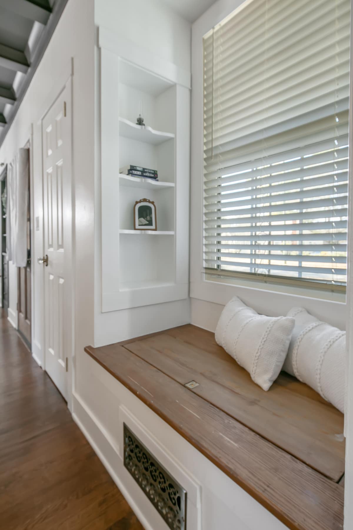 A cozy nook is highlighted by a wooden bench, adorned with two white pillows. Shelving units above display decorative items, while large windows with blinds allow natural light to illuminate the space. The wooden flooring complements the inviting atmosphere.