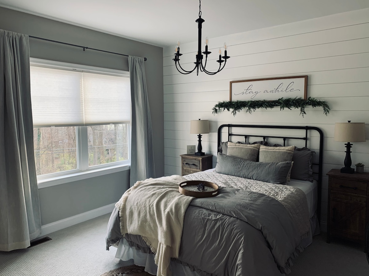 A well-appointed bedroom features a Queen size bed dressed in soft gray linens, complemented by a decorative throw. Two bedside tables with lamps frame the bed, and a furnished window invites natural light. A rustic chandelier hangs above, enhancing the room's inviting ambiance.